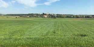 Blick auf ein grünes Feld, am Horizont ein kleiner Ort mit Kirche