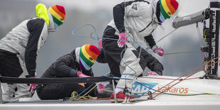 Drei Segler mit regenbogenfarbenenen Sturmhauben ackern auf einem Segelschiff