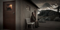 Blick auf das Bühnensetting. Eine Frau mit Maske sitzt an der Hauswand einer Hütte. Aus deren Tür schaut ein Mann mit Maske heraus