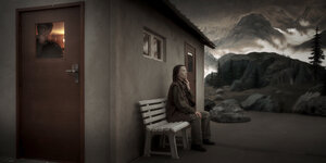 Blick auf das Bühnensetting. Eine Frau mit Maske sitzt an der Hauswand einer Hütte. Aus deren Tür schaut ein Mann mit Maske heraus