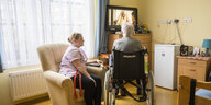 Mann im Rollstuhl vor einem Fernseher, Pflegekraft im Sessel daneben
