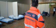 Ein Mann in orangener Johanniter-Jacke mit der Aufschrift Bevölkerungsschutz steht in einem Raum mit einfach Feldbetten