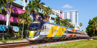 Ein bunter Zug fährt auf Schienen vor der bunten Wand einer Wohngebäudereihe in West Palm Beach in Florida, dahinter steckt ein Wohnhochhaus.