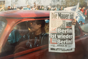 Ein Mann mit Vokuhila sitzt in einem roten Auto. Neben ihm sitzt eine Frau. Der Mann hält eine Zeitung (BZ) aus dem Auto, auf der steht: "Die Mauer ist weg! Berlin ist wieder Berlin!"