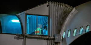 Beleuchtete Fenster eines Flugzeugs, man sieht wie ein Mann von einem anderen, der eine Schutzweste trägt in das Flugzeug geführt wird