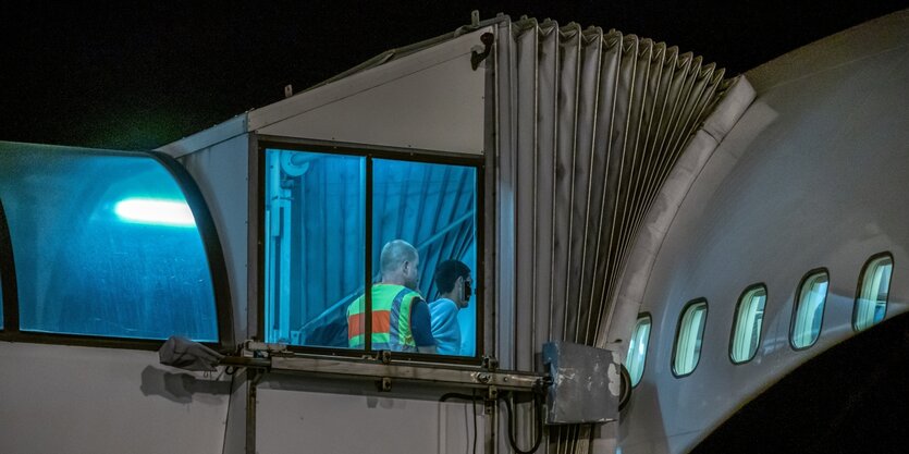 Beleuchtete Fenster eines Flugzeugs, man sieht wie ein Mann von einem anderen, der eine Schutzweste trägt in das Flugzeug geführt wird