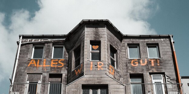 Der obere Teil eines grauen Hauses, von unten fotografiert. Auf dem Haus steht in orange "Alles wird gut"