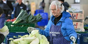 Eine Frau steht bei der Münchner Tafel mit einer Schürtze und schaut lächelnd auf Kohl