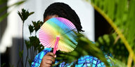 Eine Person hält sich einen Fächer in Regenbogenfarben vors Gesicht, im Hintergrund ein bogenförmiger Palmwedel