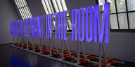 Große lilafarbene Leuchtbuchstaben in einem Ausstellungraum. Sie ergeben den Satz: There is an elephant in the room