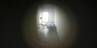 Ein Blick durch ein Türguckloch in eine karge Zelle, in der Sonnenlicht durch ein Fenster fällt. In dem Raum steht ein Bett