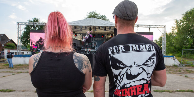 Ein Mann und eine Frau stehen auf dem Areal des „Schild und Schwert“-Festivals in einigem Abstand zu einer Bühne. Der Mann trägt ein T-Shirt mit der Aufschrift "FÜR IMMER SKINHEAD".