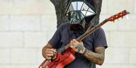 Tatowierter Straßenmusiker mit spaciger, verspiegelter Kopfhelmbedeckung spielt Gitarre