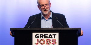 Jeremy Corbyn steht an einem Rednerpult mit einem Schild „Great Jobs“