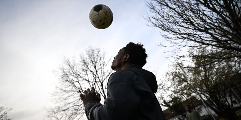 Ein Jugendlicher spielt auf einem Bolzplatz Fußball. Im Hintergrund sind kahle Baumwipfel zu sehen.