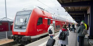 Touristen laufen mit Rollkoffern auf einen Zug der Deutschen Bahn am Gleis zu.