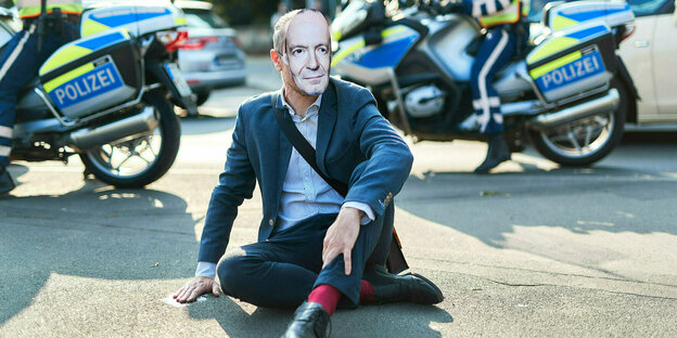 Mann sitzt aus Protest mit Maske auf einer Straße und hat seine Hand auf den Asphalt geklabt