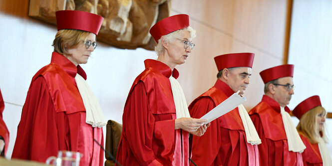 Fünf Richter*innen in roten Roben und rotem Hut stehen bei der Urteilsverkündung. Astrid Wallrabenstein liest das Urteil vor