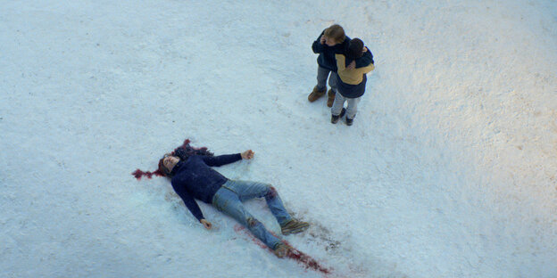Blick von oben auf einen Mann, der im Schnee liegt, mit blutigen Spuren. Zwei Personen stehen daneben