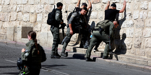 Zwei Männer stehen mit erhobenen Händen an einer Mauer und werden von Polizisten abgetastet