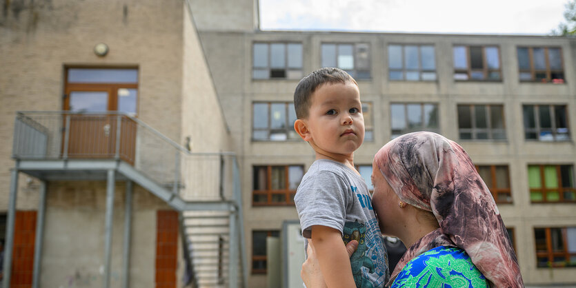 Eine Frau mit Kopftuch wendet das Gesicht ab und hebt ihren Sohn hoch, der in die Kamera schaut.