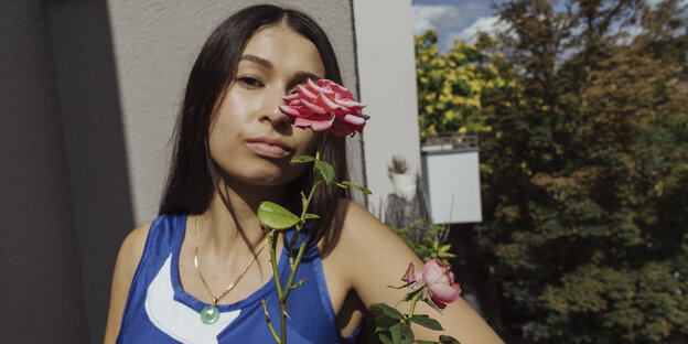 Eine junge Frau in einem blauen ärmellosen Shirt steht hinter einer Rose die ein Auge verdeckt