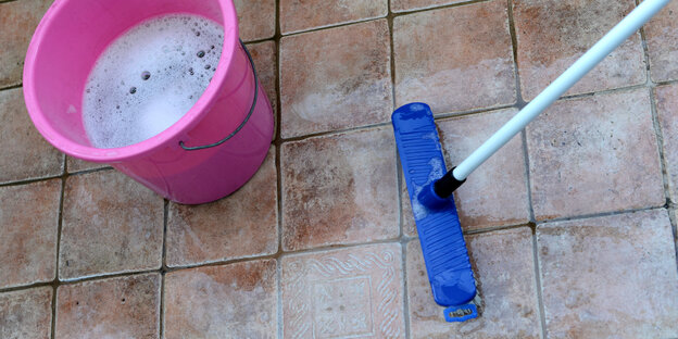 Ein pinkfarbener Eimer mit Seifenwasser und ein blauer Schrubber stehen auf einem gefliesten Boden.