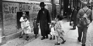 Ein Mann mit langem schwarzem Mantel und Hut führt 5 Kinder auf einem Bürgersteig, historische Schwarz-Weiß-Aufnahme