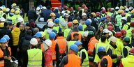 Viele Bauarbeiter mit Warnwesten und Helmen stehen in der Hamburger Hafencity, im Hintergrund ein Feuerwehrbus