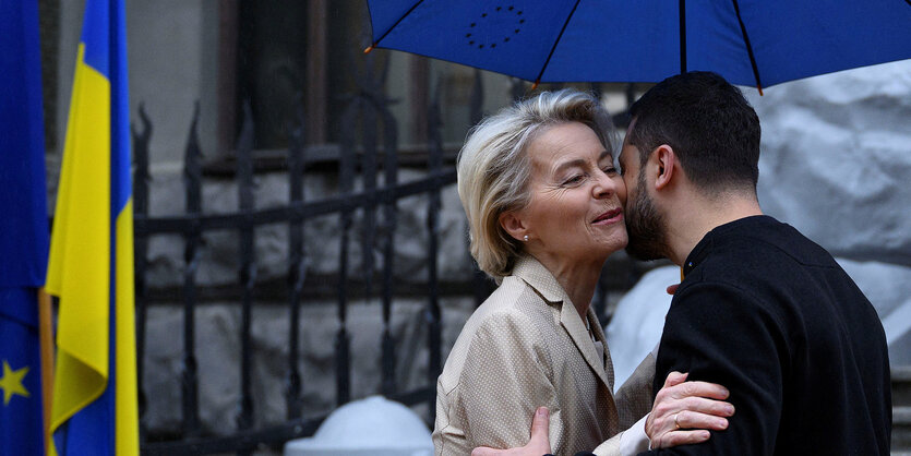 Ursula von der Leyen und Wolodymyr Selenskyj begrüßen sich unter einem blauen Regenschirm. Links im Bild ist eine ukrainische Fahne zu sehen.