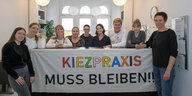 Das Team der Kiezpraxis in Kreuzberg hat sich um ein Protesttransparent versammelt, auf dem steht "Kiezpraxis muss bleiben"