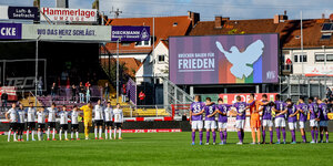 Zwei Mannschaften stehen im Osnabrücker Fußballstadion unter einer Anzeigetafel mit dem Text: "Brücken bauen für den Frieden".