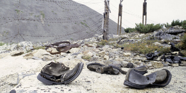 ausgetragene, kaputte Schuhe liegen auf einem Steinhaufen vor einem Stacheldrahtzaun