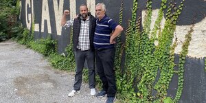Ratko Stojiljković und Irfan Beširević stehen an einer mit Efeu bewachsenen Mauer und umarmen sich. Ratko Stojiljković hebt kämpferisch die Faust