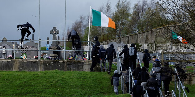 Menschen springen über den Zaun eines Friedhofs, auf dem auch eine irische Fahne steht