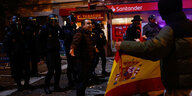 Eine Person steht auf der Straße und ruft etwas, hinter ihm Polizisten, ein anderer Mensch zeigt eine Fahne Spaniens