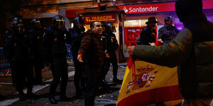 Eine Person steht auf der Straße und ruft etwas, hinter ihm Polizisten, ein anderer Mensch zeigt eine Fahne Spaniens