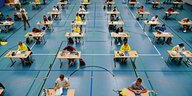 Schüler sitzen an weit auseinander gestellten Tischen in einer Turnhalle und schreiben Abitur.