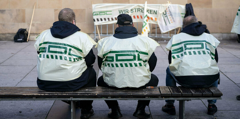 Drei Männer mit GDL-Westen sitzen auf einer Bank mit dem Rücken zu den Zuschauern