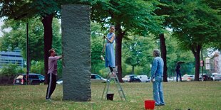 Eine Frau aif einer Leiter und ein Man putzen einen rechteckigen grauen Stein. Die Frau sprocjt m,it einem (mutmaßlichen) Passanten