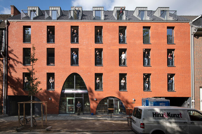 Ein viergeschossiges Haus mit Backsteinfassade, an dessen Fenstern Menschen stehen