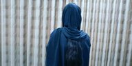 Eine Frau in blauer Burka, die Haare hängen lang über ihren Rücken, von hinten fotografiert, vor einem gemusterten Vorhang