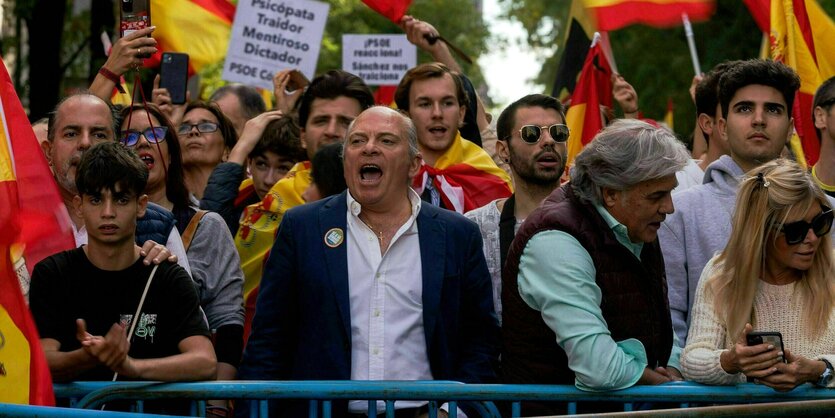 Demonstranten stehen vor einem Absperrgitter und rufen Slogans. Sie schwenken spanische Flagge oder tragen Flaggen um die Schultern