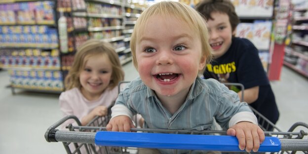 Ein Kind sitzt im Einkaufswagen, lacht und wird von zwei Freund:innen angeschoben