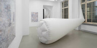 Eine riesieg aufblasbare, röhrenförmige Skulptur aus weißem PVC-Stoff ragt von der Fensterseite der Galerie ChertLüdde in den Raum. Links im Bild ist an der Wand der gerahmte Ausschnitt eines über und über mit Filzstift bemalten Stücks PVC zu sehen.