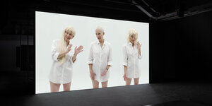 In einem dunkelen Raum steht eine Filmleinwand, auf der die selbe, blonde Frau dreimal zu sehen ist. Sie trägt ein weißes Hemd und gestikuliert