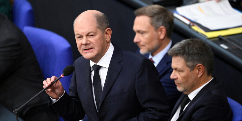 Olaf Scholz spricht im Bundestag, neben ihm sitzen mit ernsten Gesichtern Robert Habeck und Christian Lindner