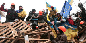 Demonstranten sitzen auf Holzpaletten, sie tragen Helme, schwingen ukrainische und europäische Flaggen und haben gute Laune