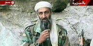 Osama Bin Laden spricht in ein Mikrofon im Camouflage-Look. Er steht vor einem Felsen, an dem ein Maschinengewehr lehnt