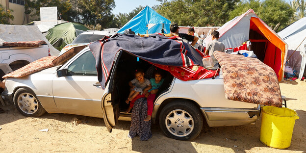 Eine Frau, deren Gesicht nicht zu erkennen ist, sitzt mit zwei Kindern auf dem Schoß auf der Rückbank eines Fahrzeugs, mit den Beinen nach draußen. Das Auto steht auf einem Sandplatz, es ist mit Habseligkeiten beladen.
